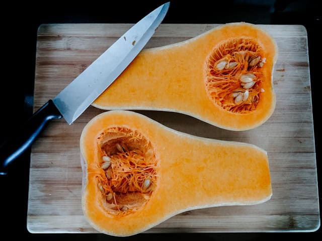 A butternut squash cut in half split open on a wooden cutting board with a knife resting on it.