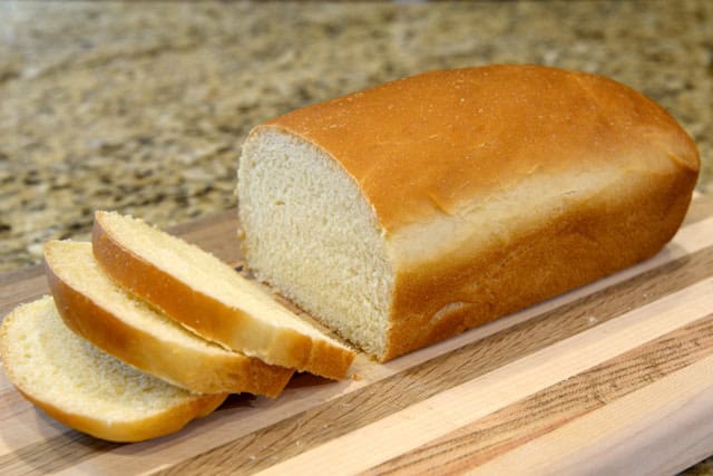A load of white bread sliced into three pieces on a wooden cutting board.