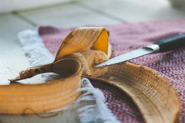 Part of a banana in its peel next to a knife on a wooden floor.