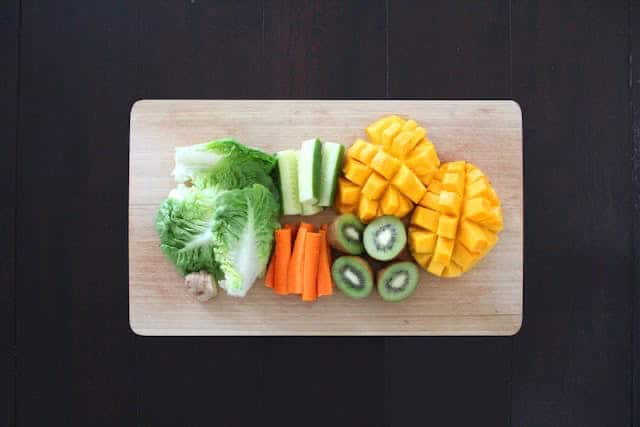 A wooden cutting board on a black surface with lettuce, celery, carrots, kiwis, and mangoes.