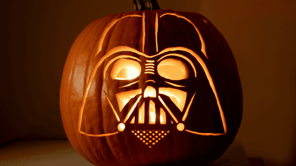 A jack-o-lantern with Darth Vader's face carved into it. The pumpkin is lit from within, casting a warm orange glow on the surrounding area. The background is a dark wall.