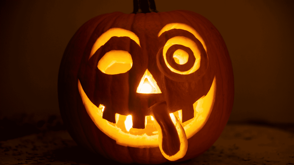 A glowing pumpkin with a silly face carved into it. The pumpkin has large, different sized eyes, simple eyebrows, and a tongue sticking out. The background is dark, and there is a candle inside the pumpkin.