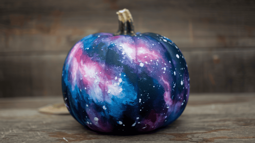 A wooden surface with a pumpkin on it. The pumpkin has been painted with a galaxy design, featuring shades of purple and blue, pink and white nebula effects, and spattered white paint for stars. The background is a wooden surface.