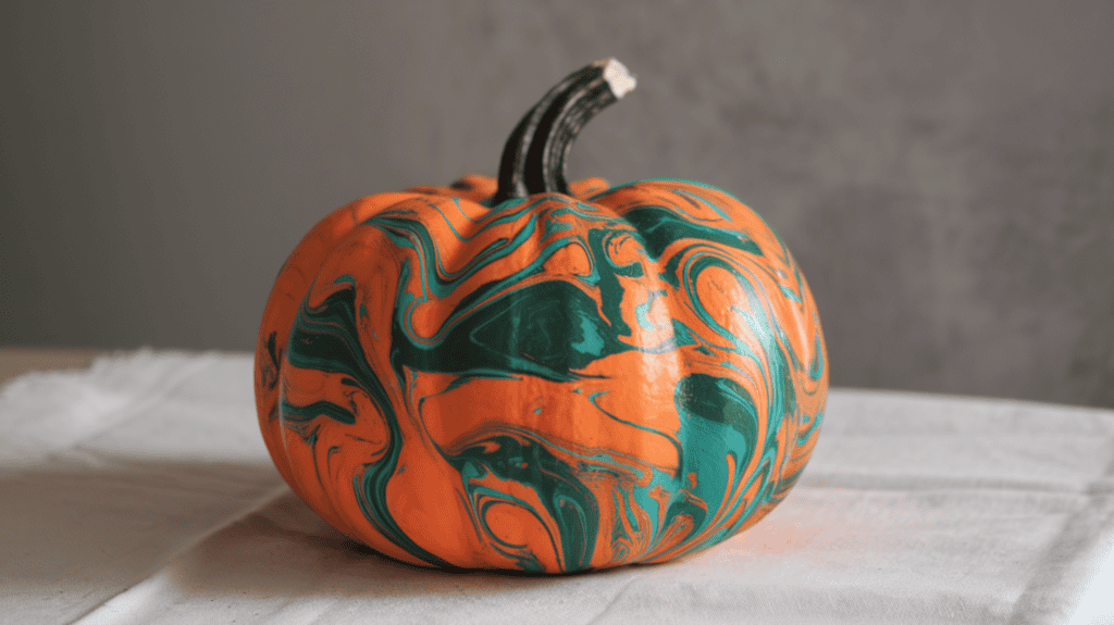 A pumpkin with a really cool marble effect. It has three different vibrant paint shades - orange, green, and blue. The orange shade is the base color, with the green and blue shades swirled throughout. The pumpkin is sitting on a table with a white cloth. The background is a gray wall.