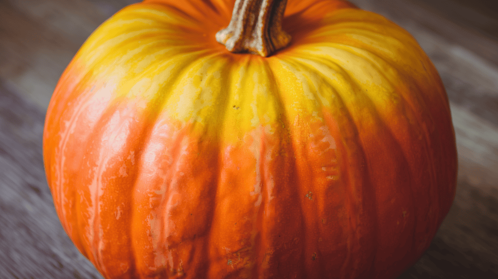 A pumpkin with an ombre effect. The pumpkin is orange with a gradient of yellows and oranges. The transitions between the colors are smooth and the overall effect is vibrant. The background is a wooden surface.