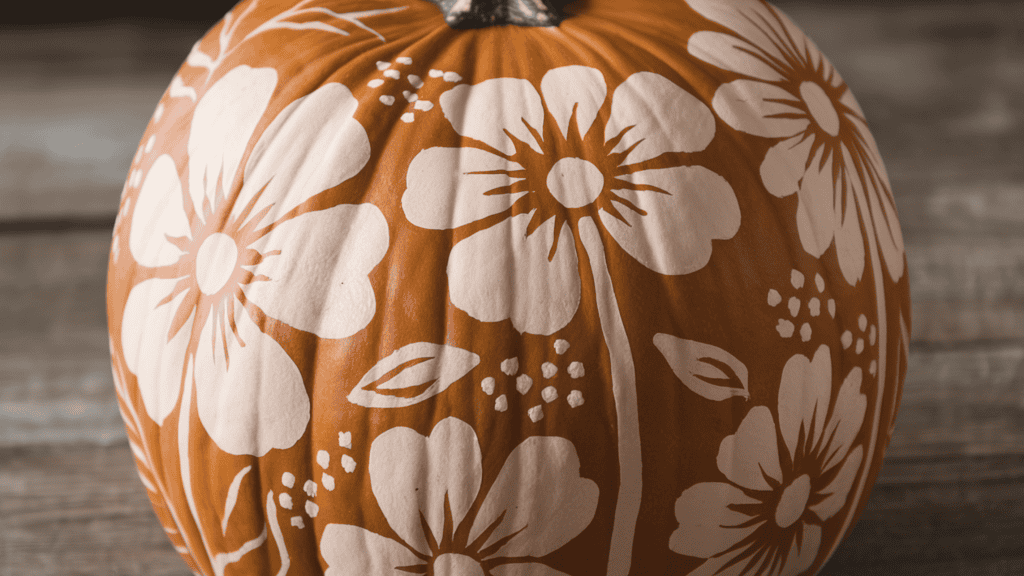 A pumpkin with a floral pattern. The pumpkin has large flower shapes painted in a lighter shade. There are basic petal definitions, leaves, and stems that are layered in. There are also tiny dots and flourishes for detail. The background is a wooden surface.