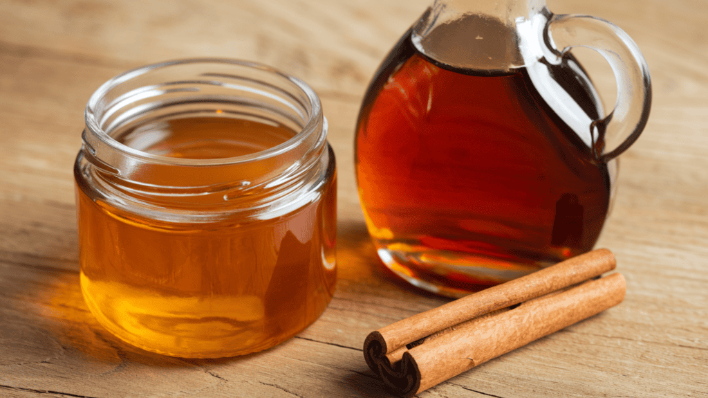 Honey and maple syrup in separate containers. The honey is in a clear jar and has a golden hue. The maple syrup is in a glass bottle with a cinnamon stick and has a dark amber color. The containers are placed on a wooden surface.