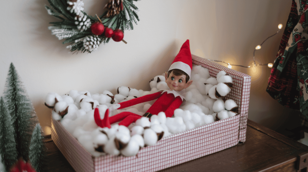 An Elf on the Shelf elf snuggled in a bed made from a tissue box with cotton ball pillows. The elf is wearing a red sweater and a red hat. There are Christmas decorations around the room, including a wreath on the wall and a tree in the corner.