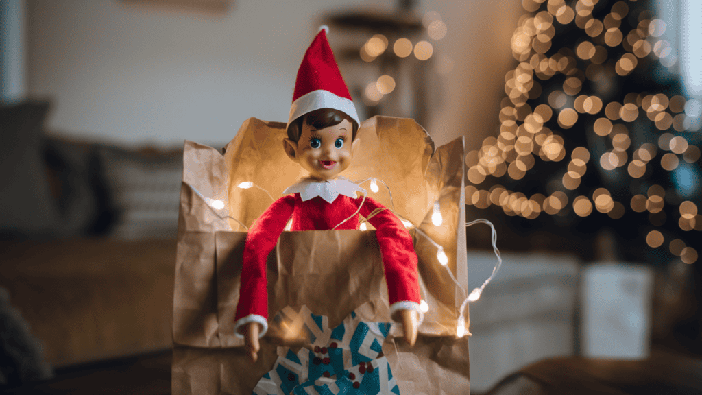An Elf on the Shelf elf emerging from a decorated paper bag. The elf is wearing a red sweater and a red hat. There are glowing fairy lights inside the bag. The background is a cozy living room with a Christmas tree.