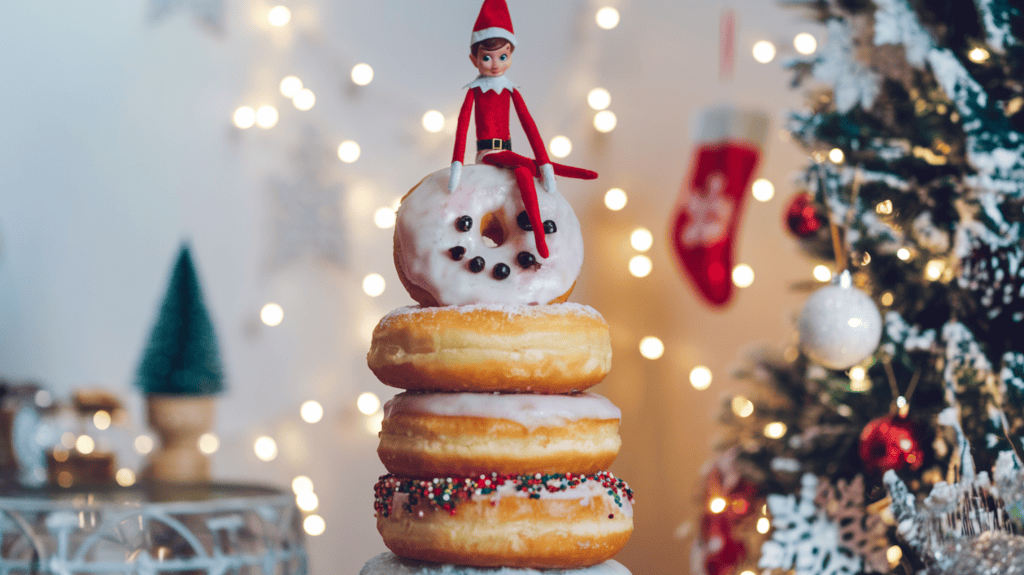 A donut snowman with an elf on the shelf elf perched on top. The snowman is made of regular-sized donuts stacked on top of each other, with a mini donut for the head. The elf is wearing a red sweater and a red hat, and is sitting on top of the snowman's head. The background is a festive setting with Christmas decorations, including a tree, ornaments, and lights.