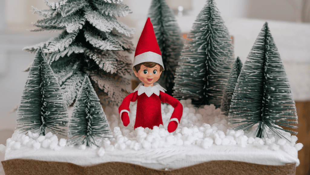 An elf on the shelf elf creating a winter wonderland scene with artificial snow and mini trees. The elf is wearing a red sweater and a red hat. The base layer is batting. The mini trees have frosted tips. The background is a white room.