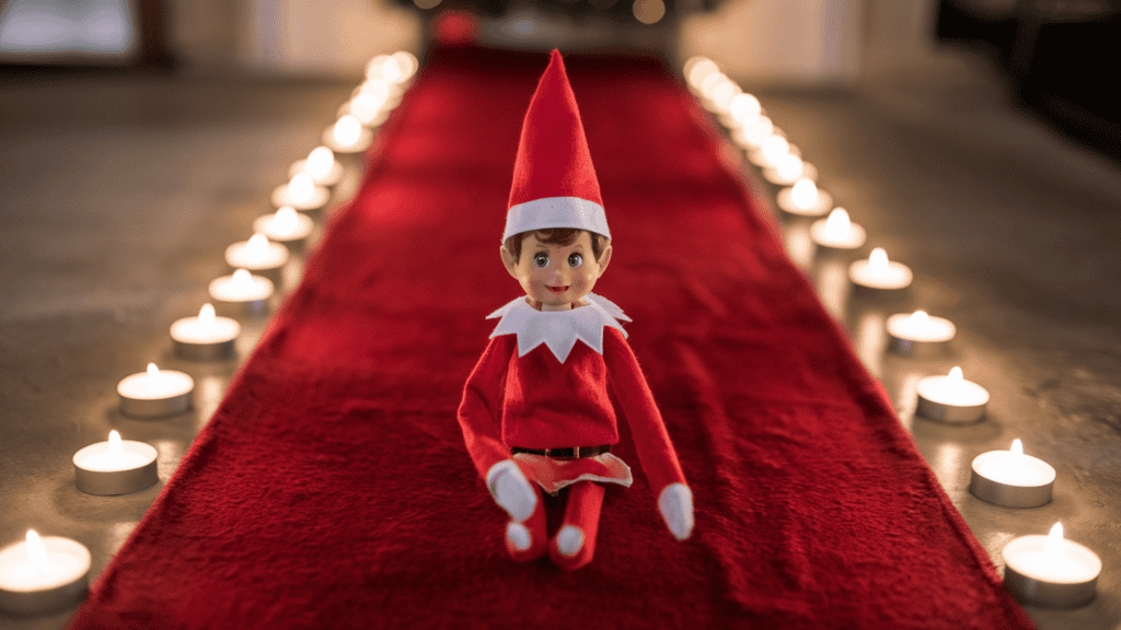 An elf on the shelf elf arriving on a red carpet entrance with battery-operated tea lights. The elf is wearing a red sweater and a red hat.