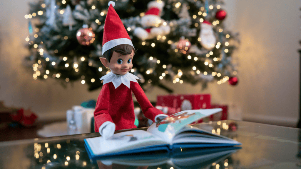 An elf on the shelf elf reading a Christmas book on the coffee table. The elf is wearing a red sweater and a red hat. The background contains a Christmas tree with ornaments and lights.