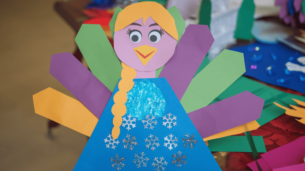 An Elsa Turkey disguise idea made of construction paper. Elsa's dress is made of blue construction paper with small snowflakes cut out from glitter paper and arranged on the dress. The background is a table with other crafts.