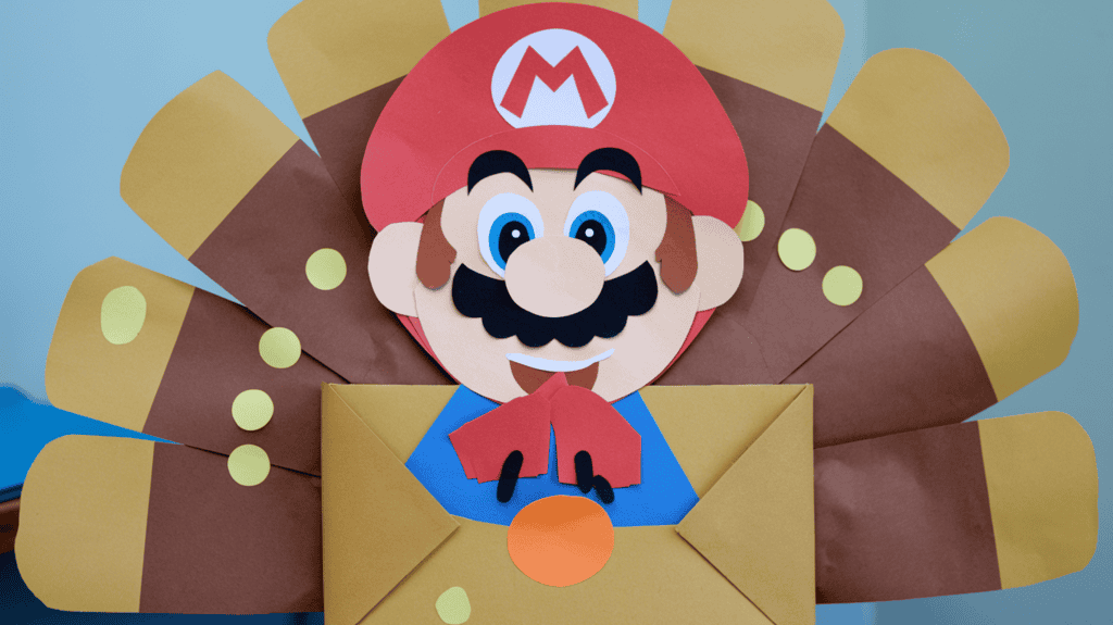 A Mario Turkey disguise idea made of construction paper. The Mario character is made out of a large red circle with a white 'M' in the middle. Mario is wearing a white hat. The turkey body is made of a brown rectangle folded in half, with a small orange circle for the head and two black lines for the eyes. There are small round stickers colored gold stuck to the craft. The background is a light blue wall.