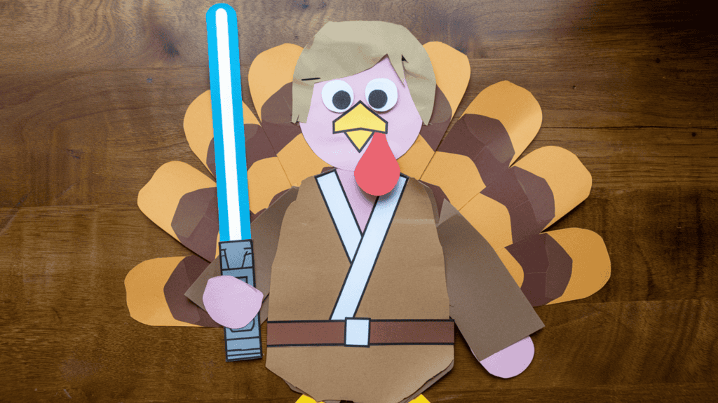 A Luke Skywalker turkey disguise idea made of construction paper. The turkey has a simple robe and is holding a lightsaber. The background is a wooden surface.