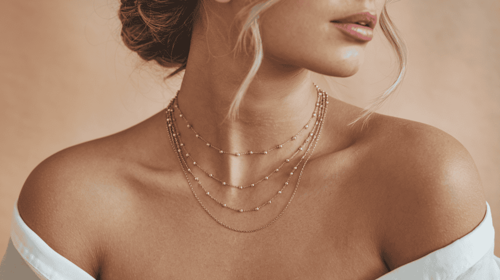 A woman wearing a gorgeous dainty layered necklace. The necklace consists of multiple gold chains of varying lengths, with small gold beads interspersed throughout. The necklace sits gracefully on the woman's collarbone. She has her hair up in a chic twist. The background is a soft beige hue.