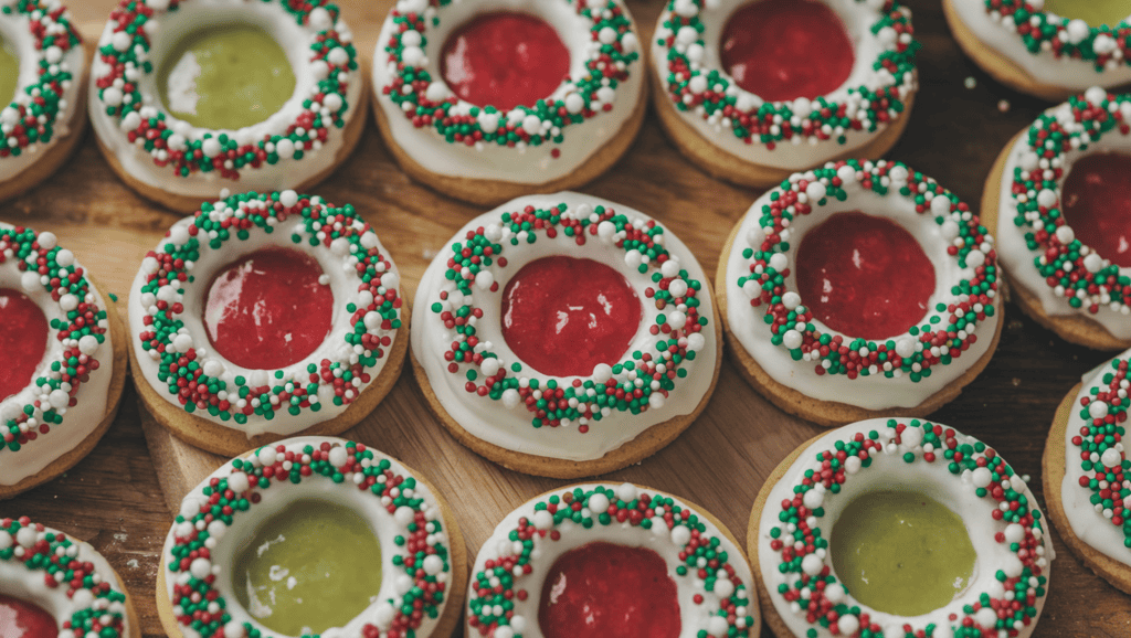Christmas-themed thumbprint cookies with festive fillings. The cookies are arranged in a circle, with each cookie having a thumbprint indentation filled with red and green jam. The cookies are adorned with sprinkles and edible glitter. The background is a wooden board.