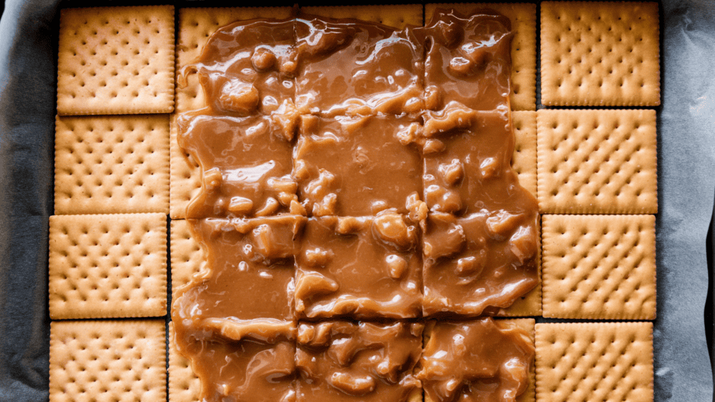 A rimmed 10x15 inch baking sheet with parchment paper and square saltine crackers arranged flat in a single layer fitting snugly together. There is a layer of toffee that has been spread evenly on top of them. The toffee has a golden-brown color and is slightly glossy. The crackers are visible through the toffee layer.