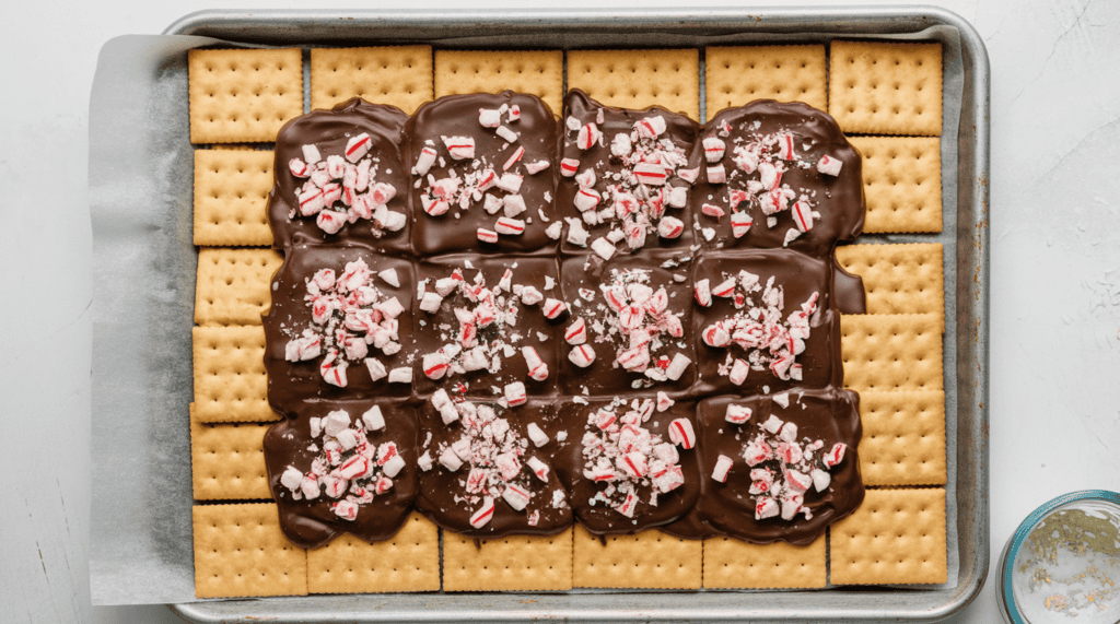 A photo of a rimmed 10x15 inch baking sheet with parchment paper and square saltine crackers arranged flat in a single layer fitting snugly together. There is a layer of chocolate that has been spread evenly on top of all the crackers with finely chopped up candy canes sprinkled across the chocolate.